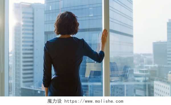 一个短头发的女商人在窗户前看室外拍的成功的女商人在一个罢工的黑色衣服在她的办公室里凝视着窗外沉思。具有个人电脑和大城市视野的现代商务办公室.